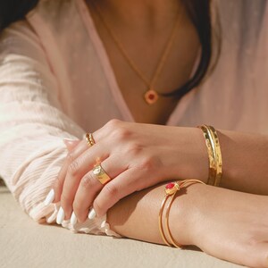 May include: A close-up of a person's hands wearing gold jewelry. The person is wearing a gold ring with a red stone, a gold bangle bracelet, and a gold chain necklace with a red stone pendant.