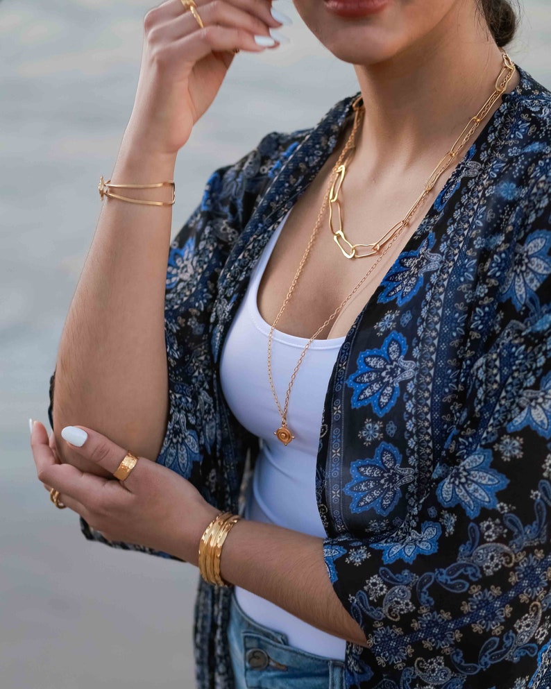 May include: A woman wearing a white tank top and a blue and black floral kimono with gold chain necklaces and bracelets.