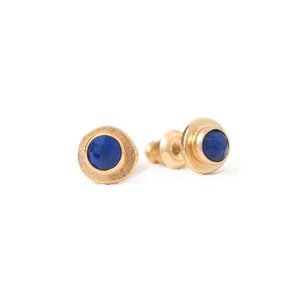Peut inclure: Une paire de boucles d'oreilles à clous dorées avec une lunette ronde et texturée. Chaque boucle d'oreille présente une pierre précieuse bleu foncé facettée. Les boucles d'oreilles sont présentées sur fond blanc.