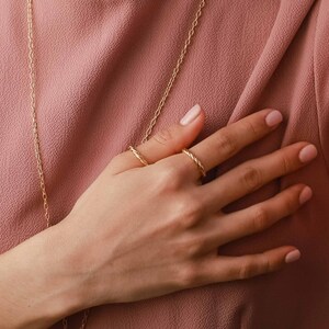 May include: Two thin gold rings with a twisted rope texture worn on the fingers. The rings feature a polished metallic finish and a continuous spiral design. A delicate gold chain necklace hangs vertically across the dusty rose fabric background.