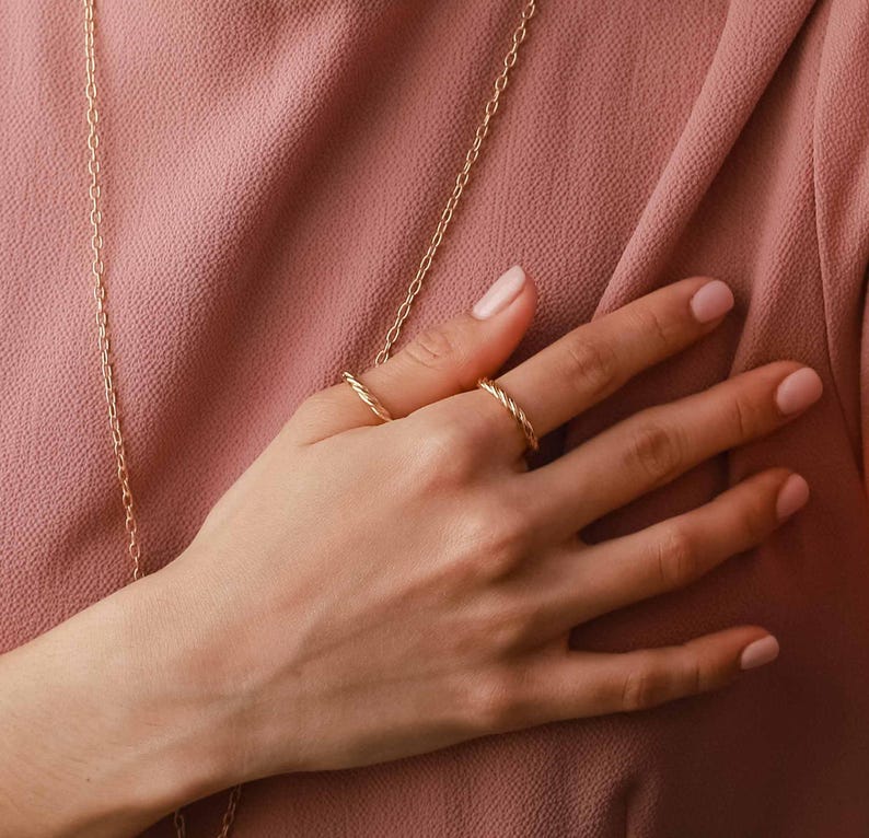 May include: Two thin gold rings with a twisted rope texture worn on the fingers. The rings feature a polished metallic finish and a continuous spiral design. A delicate gold chain necklace hangs vertically across the dusty rose fabric background.