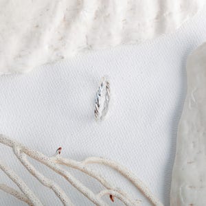May include: A silver ring featuring a twisted, rope-like texture. The band has a polished, metallic finish and a continuous, uniform design that wraps around the entire circumference of the ring, creating a subtle, spiraling pattern.
