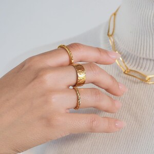 May include: Three gold-toned rings worn on fingers, including one wide band with a hammered texture and two thinner bands with a twisted rope design. A gold-toned chain link necklace is visible in the upper right corner of the frame.