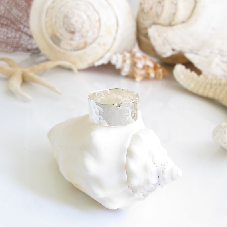 May include: A hammered silver band ring, sitting on a white seashell. The ring is wide and has a textured surface.