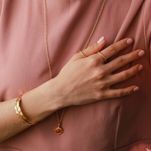 May include: A gold necklace with a red gemstone pendant, a gold bracelet, and two gold rings on a hand wearing a pink shirt.