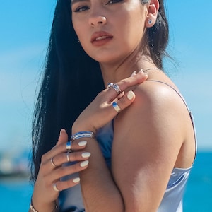 May include: A woman with long black hair wearing a light blue satin top and several silver rings on her fingers. She is wearing a silver bracelet on her wrist and a small silver earring in her left ear.