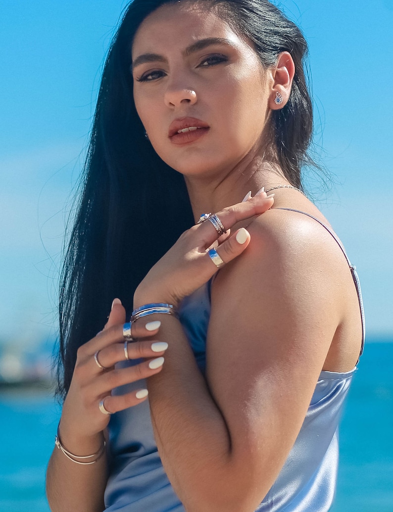 May include: A woman with long black hair wearing a light blue satin top and several silver rings on her fingers. She is wearing a silver bracelet on her wrist and a small silver earring in her left ear.