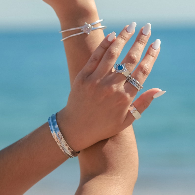 May include: A woman's hand wearing a silver ring with a blue stone and several other silver rings. The hand is also wearing a silver bracelet with a textured design.