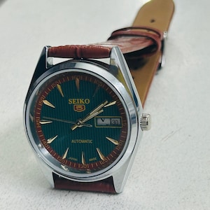 May include: A Seiko 5 automatic watch with a green dial and gold-colored markers. The watch has a silver-colored case and a brown leather strap. The date is displayed at the 3 o'clock position. The watch face has the text "SEIKO 5 AUTOMATIC JAPAN MADE".