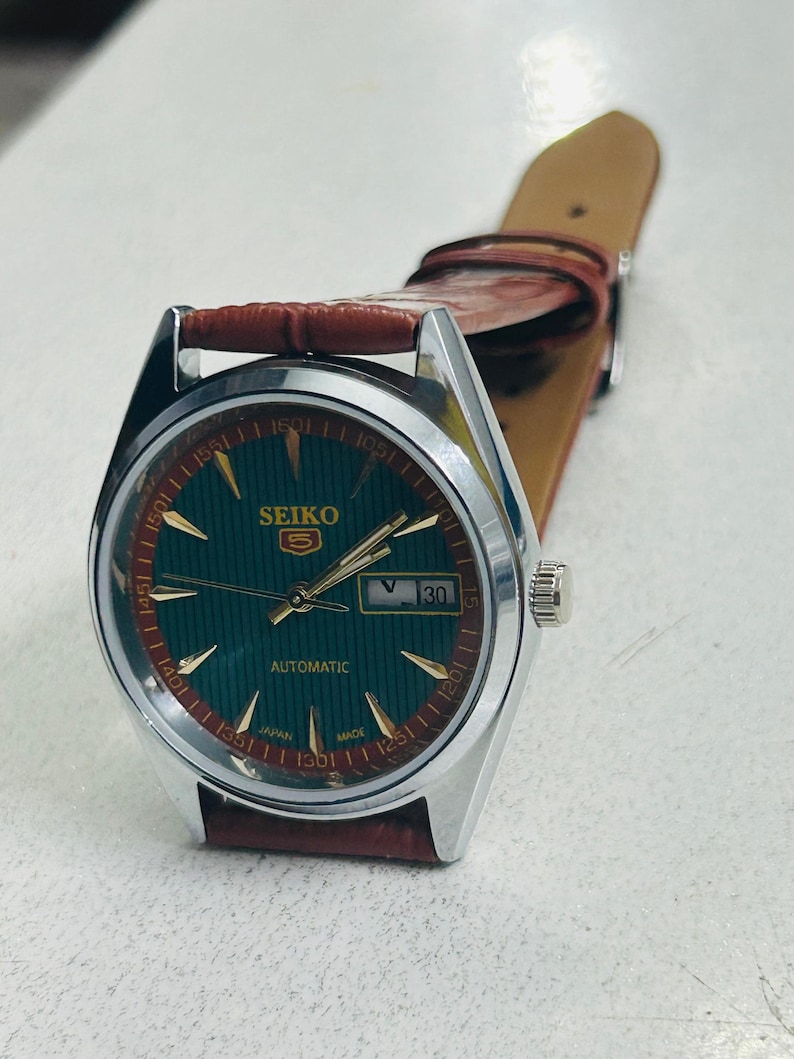 May include: A Seiko 5 automatic watch with a green dial and gold-colored markers. The watch has a silver-colored case and a brown leather strap. The date is displayed at the 3 o'clock position. The watch face has the text "SEIKO 5 AUTOMATIC JAPAN MADE".