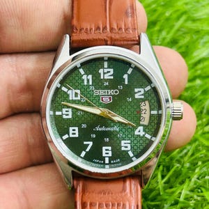 May include: A silver-toned Seiko automatic watch with a green dial and a brown leather strap. The watch face features white numerals, a date display, and gold-toned hands. The strap is a textured brown leather.