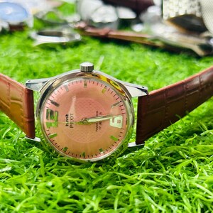 May include: A vintage HMT Pilot watch with a salmon-coloured dial and a brown leather strap. The watch has a silver-coloured case and features the date at the 3 o'clock position. The watch is displayed on green grass.