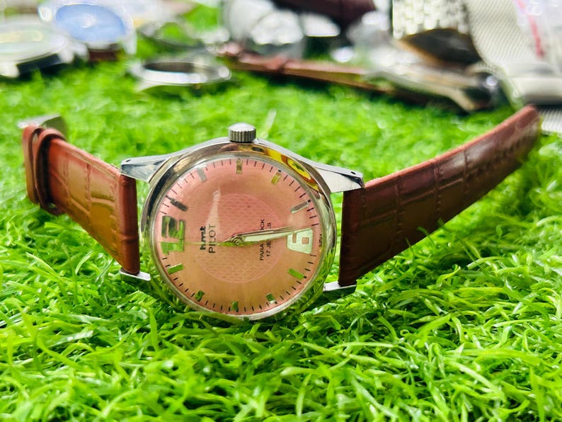 May include: A vintage HMT Pilot watch with a salmon-coloured dial and a brown leather strap. The watch has a silver-coloured case and features the date at the 3 o'clock position. The watch is displayed on green grass.