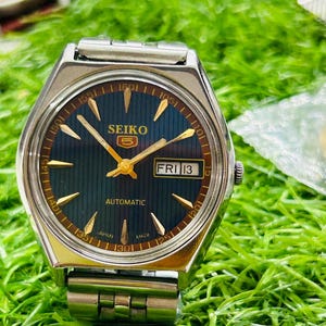 May include: A silver-toned Seiko 5 automatic wristwatch with a dark blue face and gold-colored hands and markers. The watch displays "FRI 13" in a small window. The watchband is silver and the watch is set against a green background.
