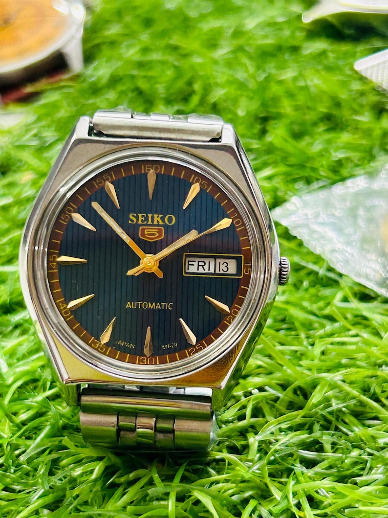 May include: A silver-toned Seiko 5 automatic wristwatch with a dark blue face and gold-colored hands and markers. The watch displays "FRI 13" in a small window. The watchband is silver and the watch is set against a green background.