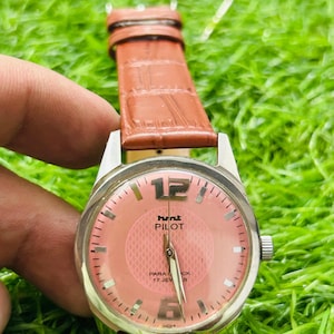 May include: A vintage wristwatch with a salmon-coloured face and a brown leather strap. The watch face features the text "hunt PILOT" and "PARA CK 17 JEW S". The watch has silver-coloured metal casing.