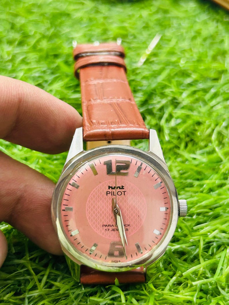 May include: A vintage wristwatch with a salmon-coloured face and a brown leather strap. The watch face features the text "hunt PILOT" and "PARA CK 17 JEW S". The watch has silver-coloured metal casing.