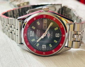 May include: A Citizen automatic wristwatch with a silver band and a black dial with red accents. The watch face displays the day and date, with the text "CITIZEN AUTOMATIC" and "21 JEWELS". The watch is made in Japan.