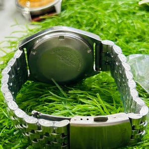 May include: A silver-toned wristwatch with a stainless steel band is displayed. The watch face is not visible, but the back casing shows text. The band is a linked style with a clasp. Another watch with a brown face is visible in the background.