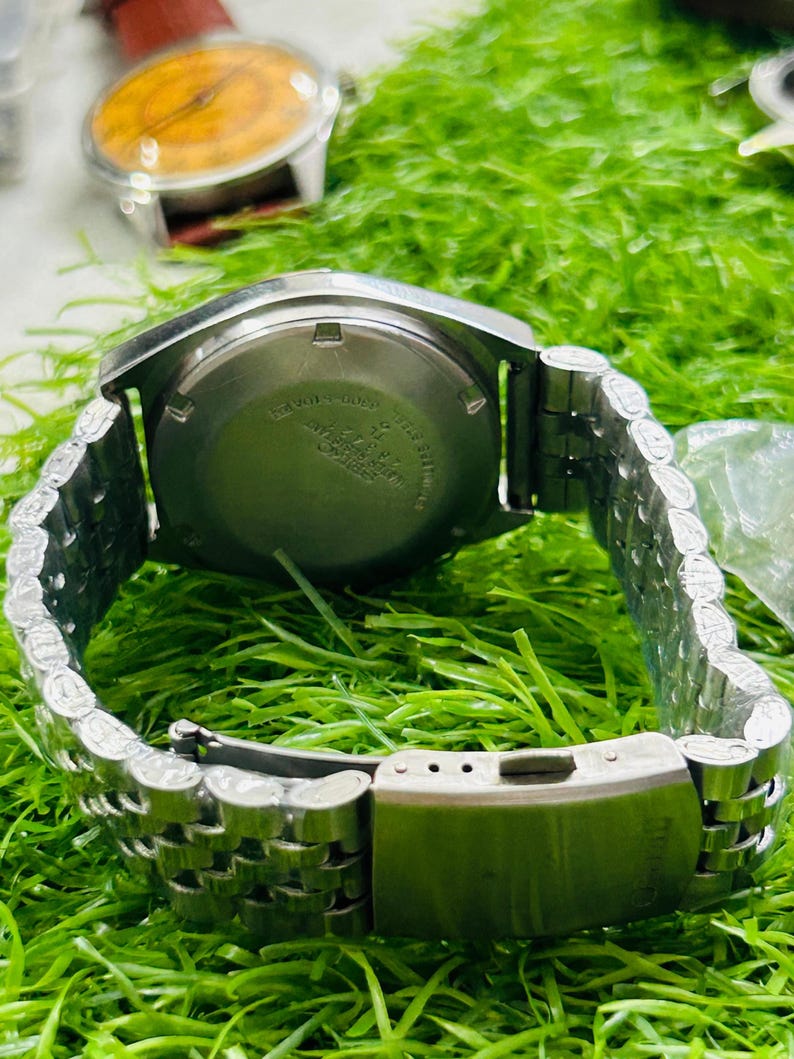 May include: A silver-toned wristwatch with a stainless steel band is displayed. The watch face is not visible, but the back casing shows text. The band is a linked style with a clasp. Another watch with a brown face is visible in the background.