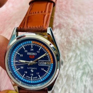 May include: A Seiko automatic wristwatch with a blue dial and a brown leather strap. The watch face displays the day and date, with the text "SAT 24" visible. The watch has a silver-colored case and a brown leather band.