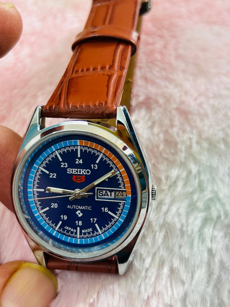May include: A Seiko automatic wristwatch with a blue dial and a brown leather strap. The watch face displays the day and date, with the text "SAT 24" visible. The watch has a silver-colored case and a brown leather band.