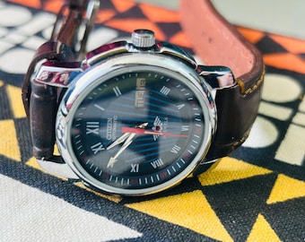 May include: A Citizen automatic wristwatch with a black dial, silver case, and brown leather strap. The watch face displays Roman numerals, day and date indicators, and red accents. The watch is set against a patterned background.