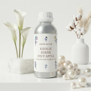 May include: A silver bottle of Kayalie Edenn Juicy Apple concentrated perfume oil. The label features the brand name and product details, with floral accents. White calla lilies and decorative pearls are also in the image.