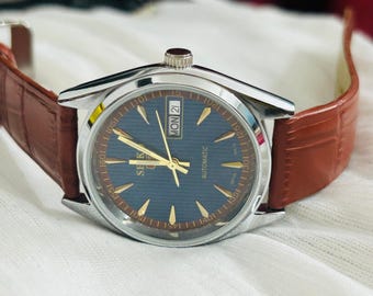 May include: A silver-toned wristwatch with a dark blue face, gold hands and markers, and a brown leather band. The watch face displays the day and date. The band has a textured, crocodile-like pattern.