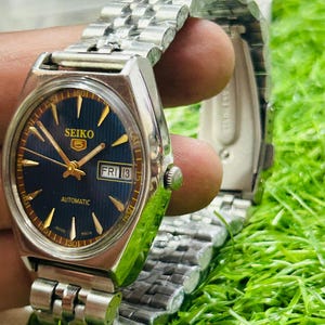 May include: A vintage Seiko 5 automatic wristwatch with a silver stainless steel band. The watch face is a deep blue with gold-colored hands and markers. The day and date are displayed at the 3 o'clock position. The watch is shown on a green surface.