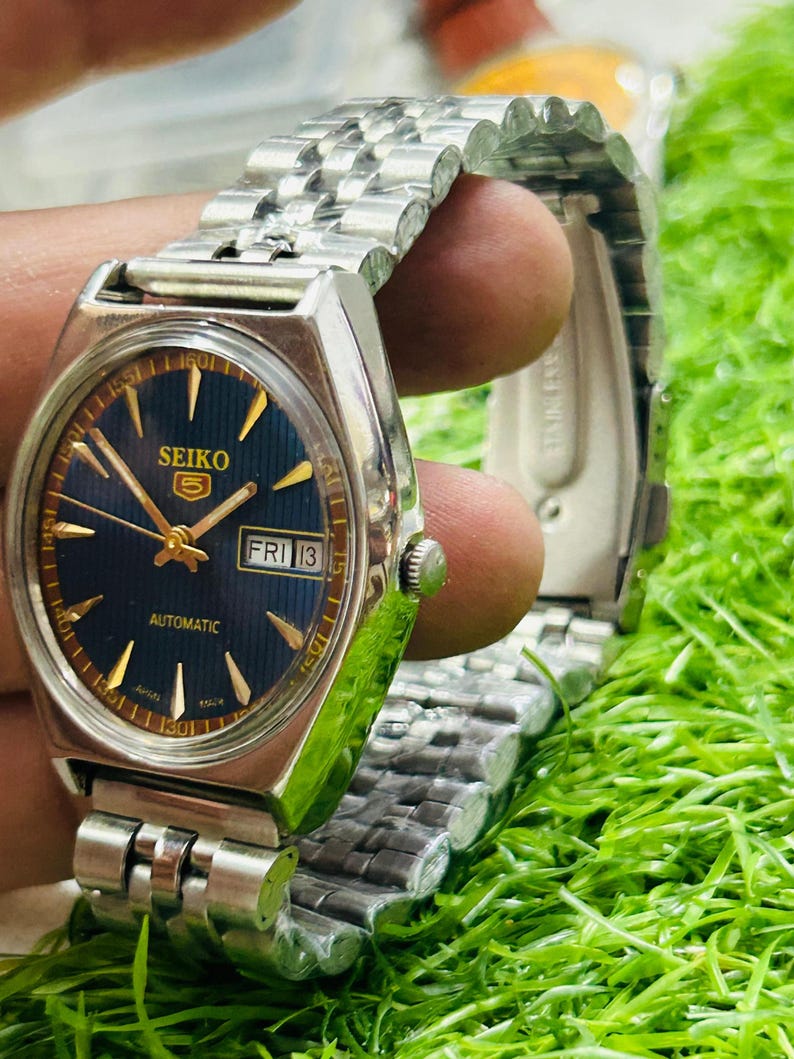 May include: A vintage Seiko 5 automatic wristwatch with a silver stainless steel band. The watch face is a deep blue with gold-colored hands and markers. The day and date are displayed at the 3 o'clock position. The watch is shown on a green surface.