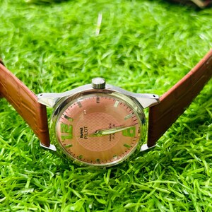 May include: A vintage wristwatch with a silver case and a brown leather strap. The watch face is salmon-coloured with green numerals and hands. The watch is labelled "Time Pilot" and "27 Jewels".