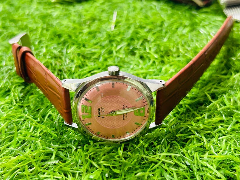 May include: A vintage wristwatch with a silver case and a brown leather strap. The watch face is salmon-coloured with green numerals and hands. The watch is labelled "Time Pilot" and "27 Jewels".