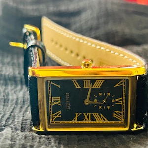 May include: A rectangular Seiko watch with a gold-tone case and black face. The watch features gold Roman numerals and hands, with a small seconds dial. The watch has a black leather band and a gold buckle.