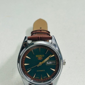 May include: A silver-toned Seiko 5 automatic watch with a green dial and gold-colored hands and markers. The watch has a brown leather strap and a date display at the 3 o'clock position. The watch face has the text "SEIKO 5 AUTOMATIC".