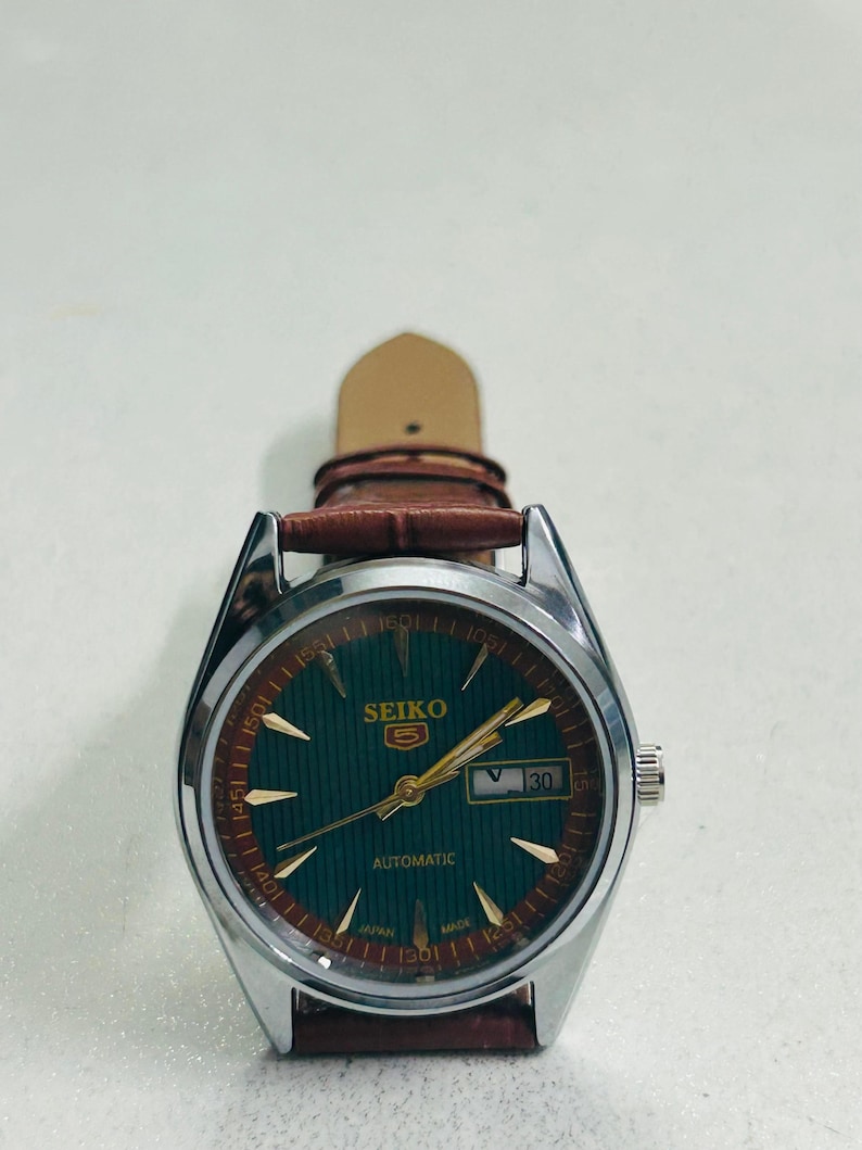 May include: A silver-toned Seiko 5 automatic watch with a green dial and gold-colored hands and markers. The watch has a brown leather strap and a date display at the 3 o'clock position. The watch face has the text "SEIKO 5 AUTOMATIC".