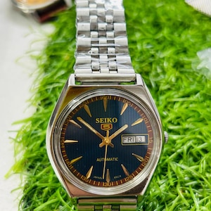 May include: A vintage Seiko 5 automatic watch with a silver stainless steel strap. The watch face is navy blue with gold hands and markers. The day and date are displayed at the 3 o'clock position. The watch is set against a green background.