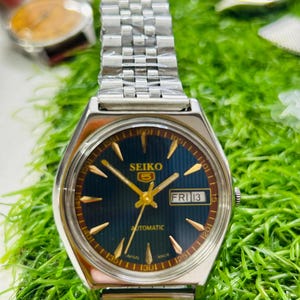 Op de afbeelding: Een Seiko 5 automatisch horloge met een zilveren band en een donkerblauwe wijzerplaat. De wijzerplaat toont "FRI 13" en goudkleurige wijzers en markeringen. Het horloge staat tegen een groene achtergrond.