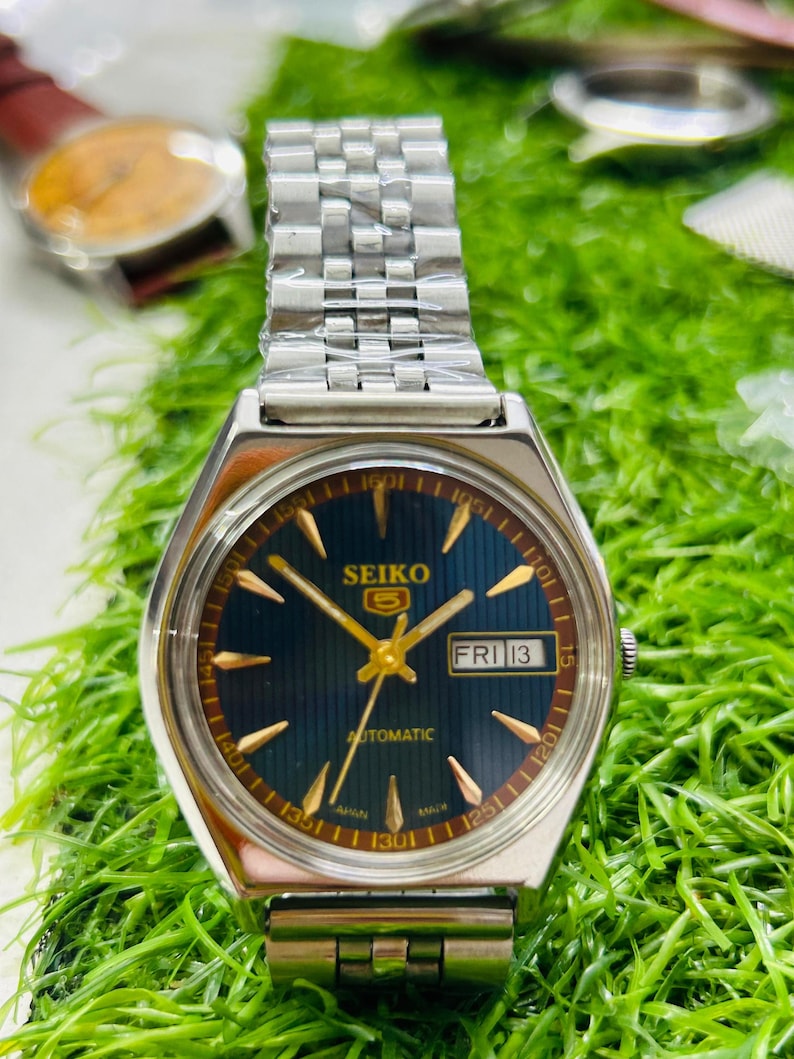 May include: A Seiko 5 automatic watch with a silver band and a dark blue face. The watch face displays the day as "FRI 13" and gold-colored hands and markers. The watch is set against a green background.