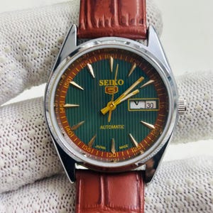 May include: A vintage Seiko automatic wristwatch with a green dial and a brown leather strap. The watch face features gold hands and markers, with the date displayed at the 3 o'clock position. The watch is marked "SEIKO" and "AUTOMATIC".