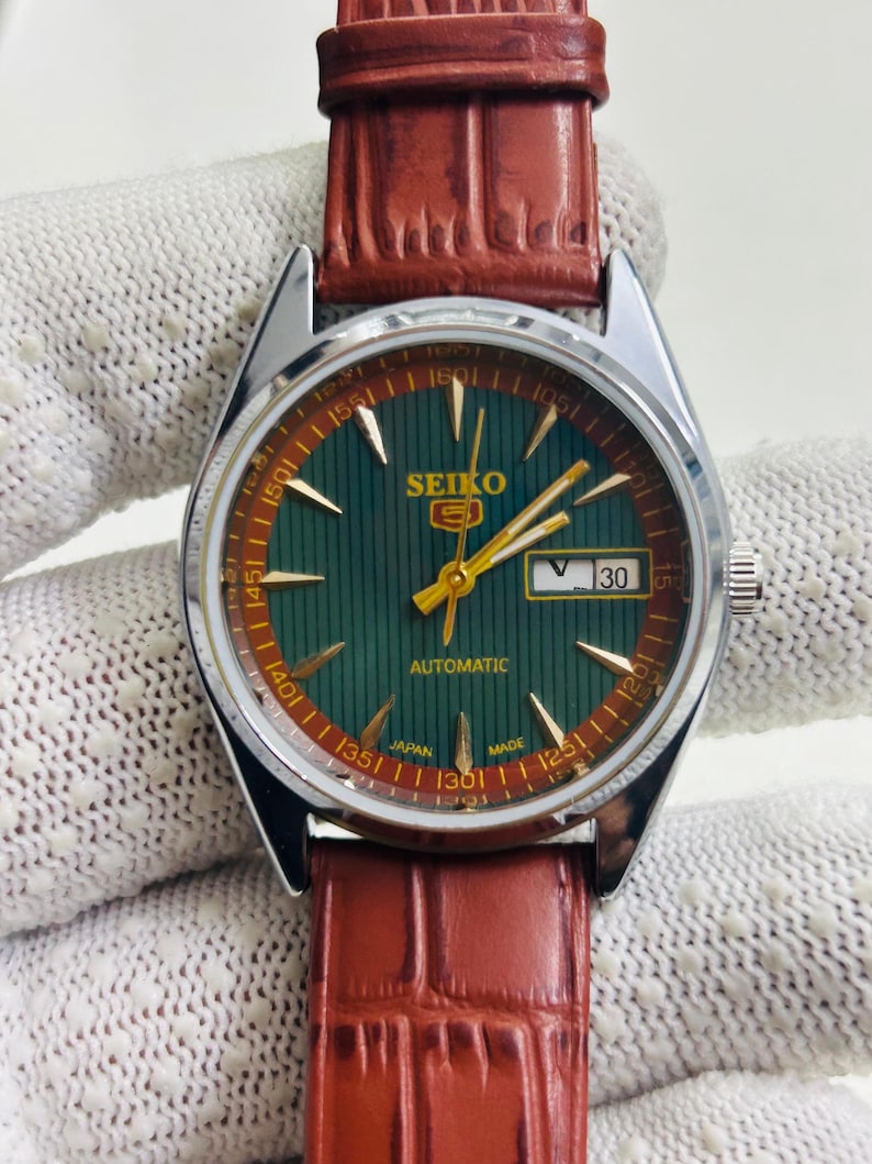 May include: A vintage Seiko automatic wristwatch with a green dial and a brown leather strap. The watch face features gold hands and markers, with the date displayed at the 3 o'clock position. The watch is marked "SEIKO" and "AUTOMATIC".