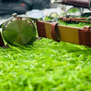 May include: A silver wristwatch with a brown leather strap rests on green grass. The watch face is visible, and the strap is secured with a silver buckle. The watch has a classic design, suitable for any occasion.