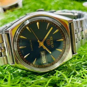 May include: A silver-toned Seiko 5 automatic wristwatch with a dark green face, gold hands and markers, and a date display. The watch has a silver band and is set against a green background.