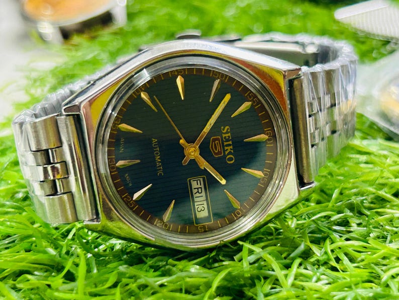 May include: A silver-toned Seiko 5 automatic wristwatch with a dark green face, gold hands and markers, and a date display. The watch has a silver band and is set against a green background.