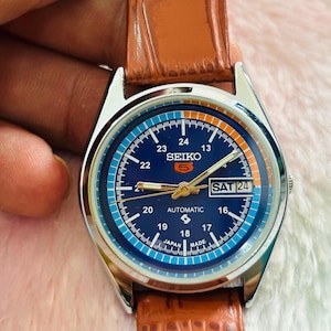 May include: A Seiko automatic wristwatch with a blue dial and a brown leather strap. The watch face displays the day and date, with the text "SEIKO 5 AUTOMATIC" visible. The watch has a silver-colored case and a brown leather band.