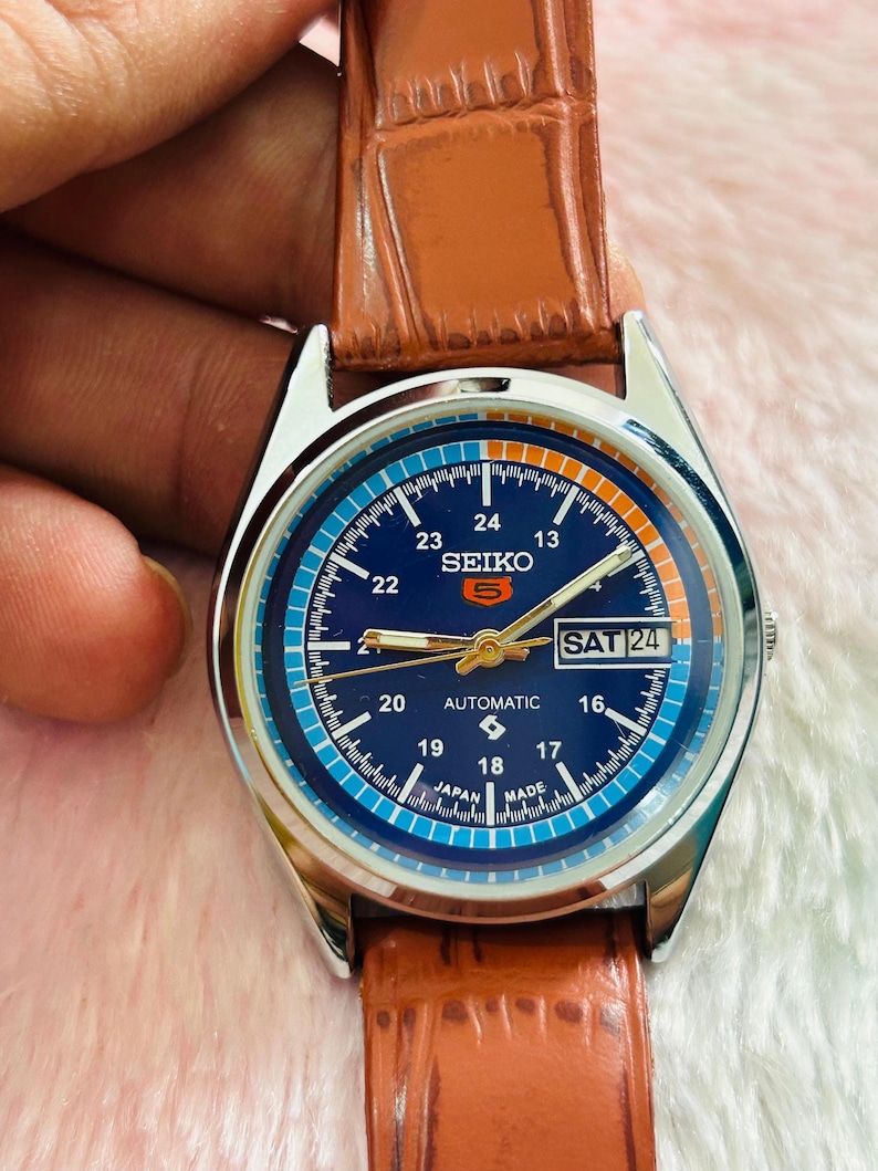 May include: A Seiko automatic wristwatch with a blue dial and a brown leather strap. The watch face displays the day and date, with the text "SEIKO 5 AUTOMATIC" visible. The watch has a silver-colored case and a brown leather band.