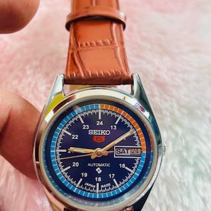 May include: A Seiko 5 automatic watch with a blue dial and a brown leather strap. The watch face displays the day and date, with the day reading "SAT 24". The watch has a silver-colored case.