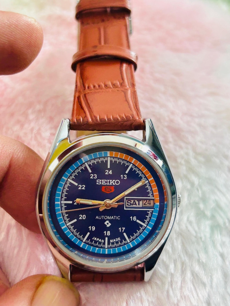 May include: A Seiko 5 automatic watch with a blue dial and a brown leather strap. The watch face displays the day and date, with the day reading "SAT 24". The watch has a silver-colored case.