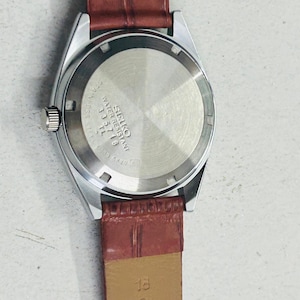 May include: A silver-toned Seiko watch with a brown leather band. The watch face is not visible, but the back of the watch shows the Seiko logo and model information. The band is a reddish-brown color.