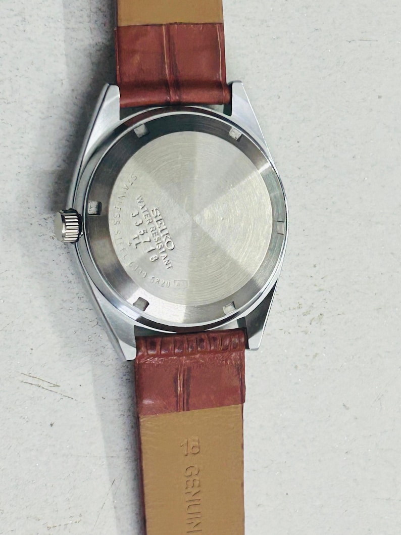 May include: A silver-toned Seiko watch with a brown leather band. The watch face is not visible, but the back of the watch shows the Seiko logo and model information. The band is a reddish-brown color.